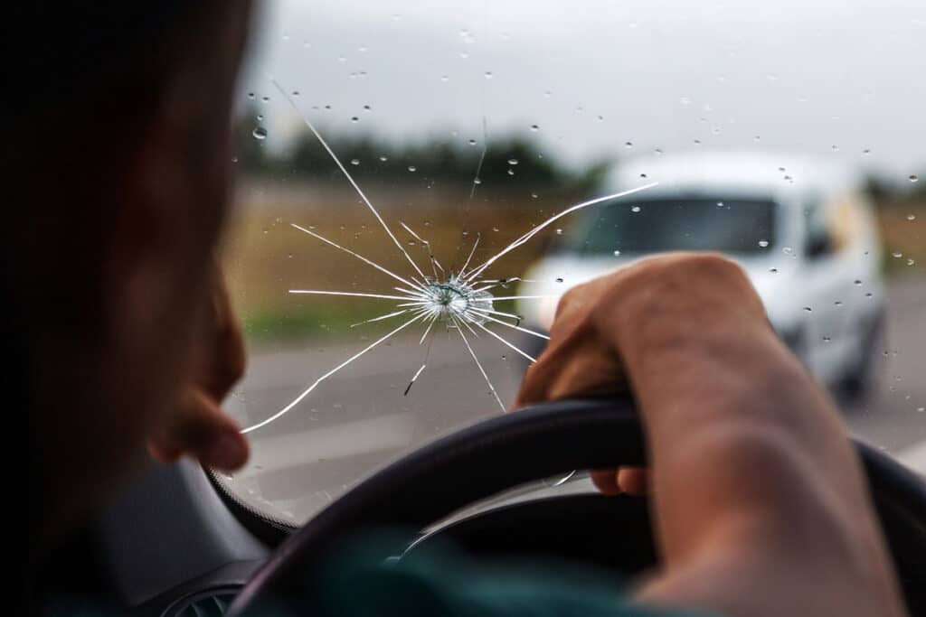 A close-up view of a cracked windshield with a hand gripping the steering wheel; a white vehicle is visible on the road ahead, and raindrops are on the glass.