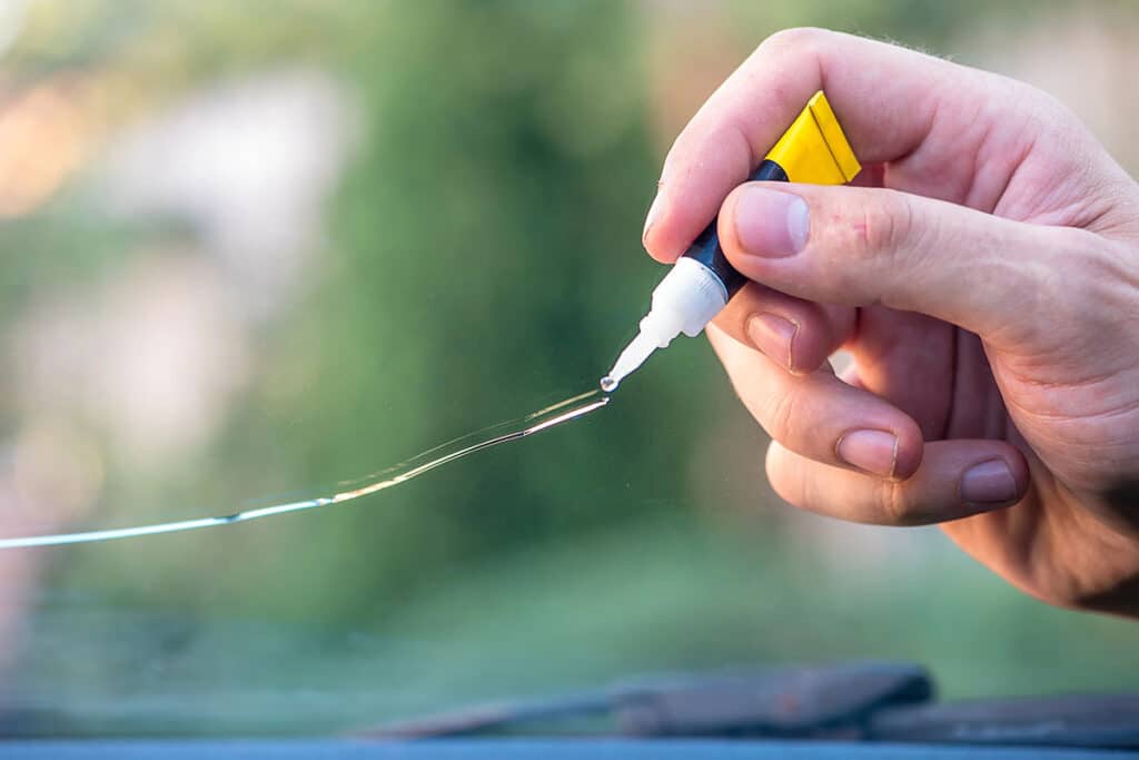 A hand holding a small bottle of adhesive, with a drop of glue forming at the tip, poised to apply it to a crack in a piece of glass.