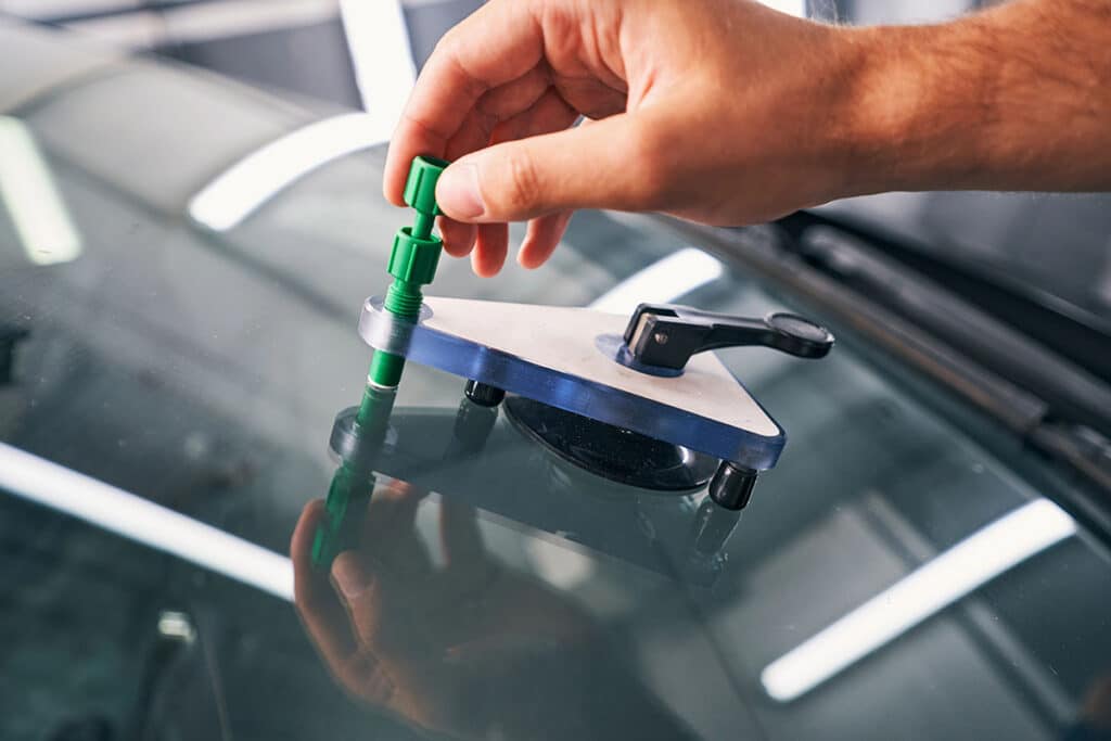 A close-up of a hand adjusting a green tool on a suction cup apparatus placed on a car windshield, highlighting the repair process for a windshield chip or crack.