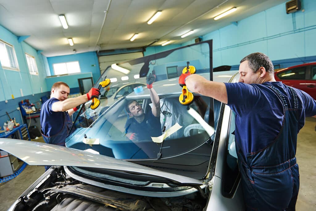Two auto technicians are installing a windshield on a car in a workshop. One technician is using suction cups to lift the windshield, while the other assists by positioning it. The workshop has blue walls and various tools and vehicles in the background.
