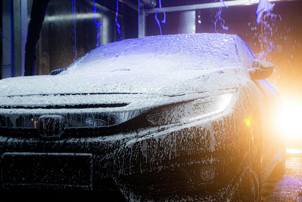 A black car covered in foam is shown in a car wash, illuminated by colorful lights with water droplets cascading down its surface.